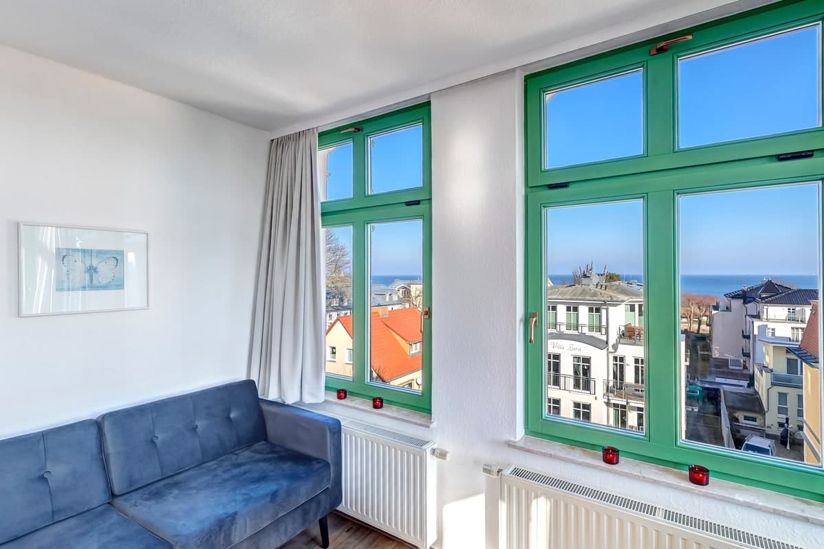 Wohnzimmer mit blauem Sofa und grünen Sprossenfenstern, dahinter Dächer und die Ostsee – als einzelnes Foto