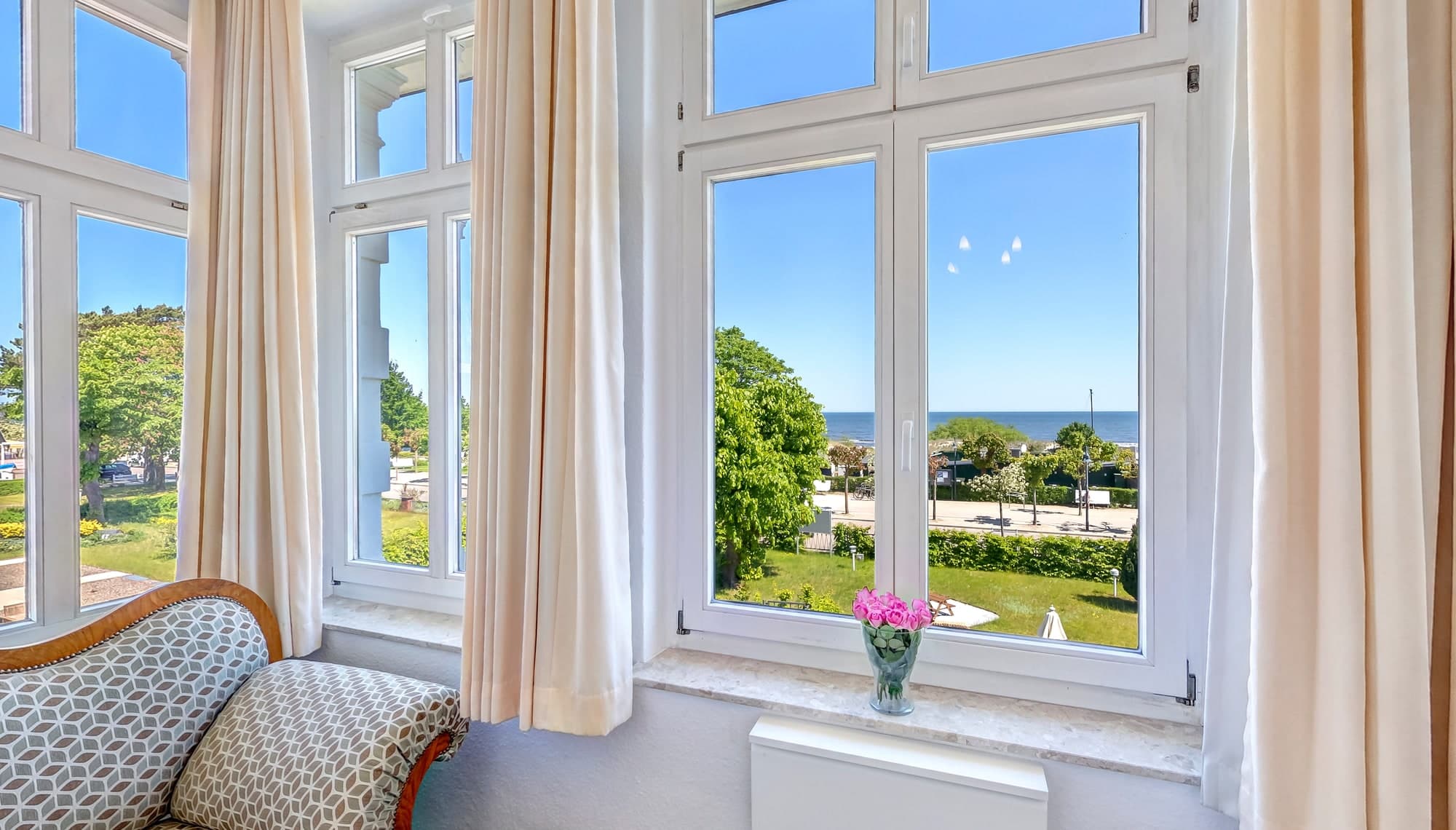 Erker mit hellen Vorhängen, Sofa und Rosen auf der Fensterbank – Blick über Garten und Strandpromenade auf die Ostsee