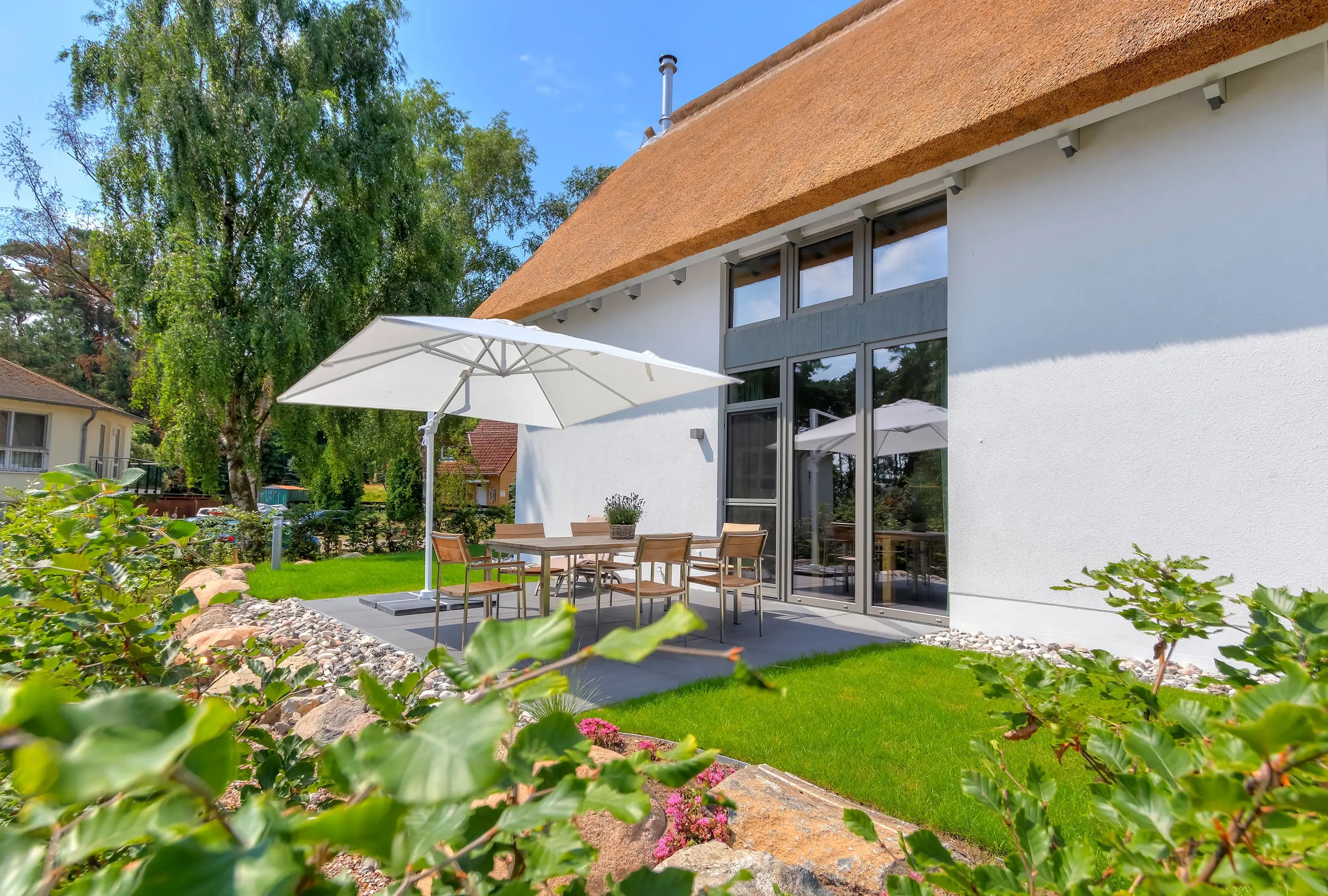 Reetgedecktes Haus mit großer Fensterfront, Terrasse und Sonnenschirm im Garten
