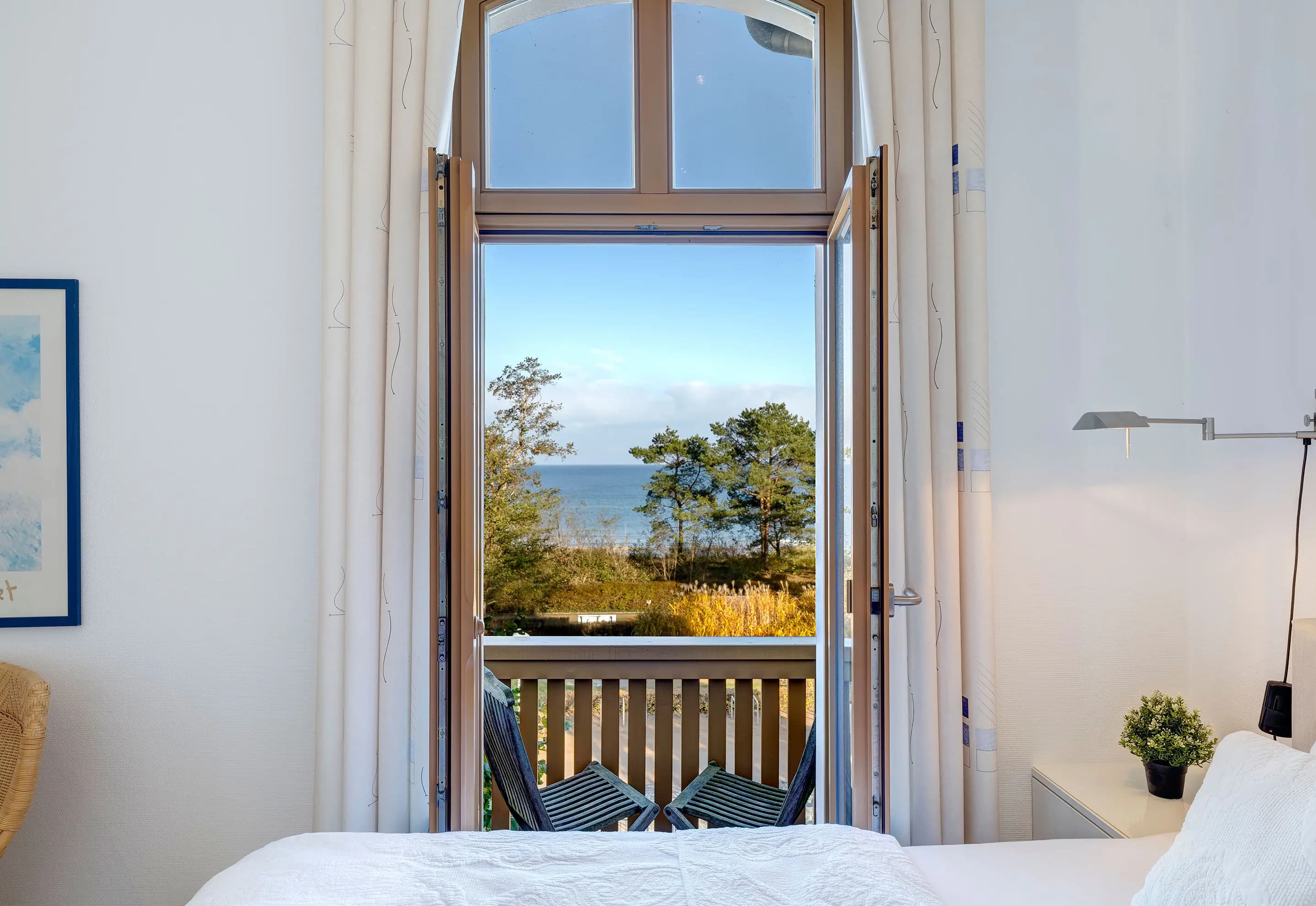 Helles Schlafzimmer mit offener Balkontür und Blick über die Kiefern auf die Ostsee